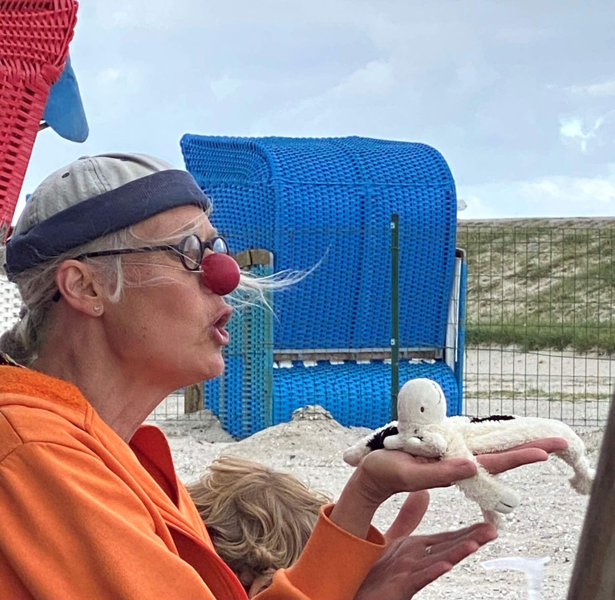 Eine Frau mit Clownsnase erzählt eine Geschichte. In ihrer Hand hält sie ein Stoffschaf. Im Hintergrund sind ein blauer Strandkorb und die Dünen zu sehen.