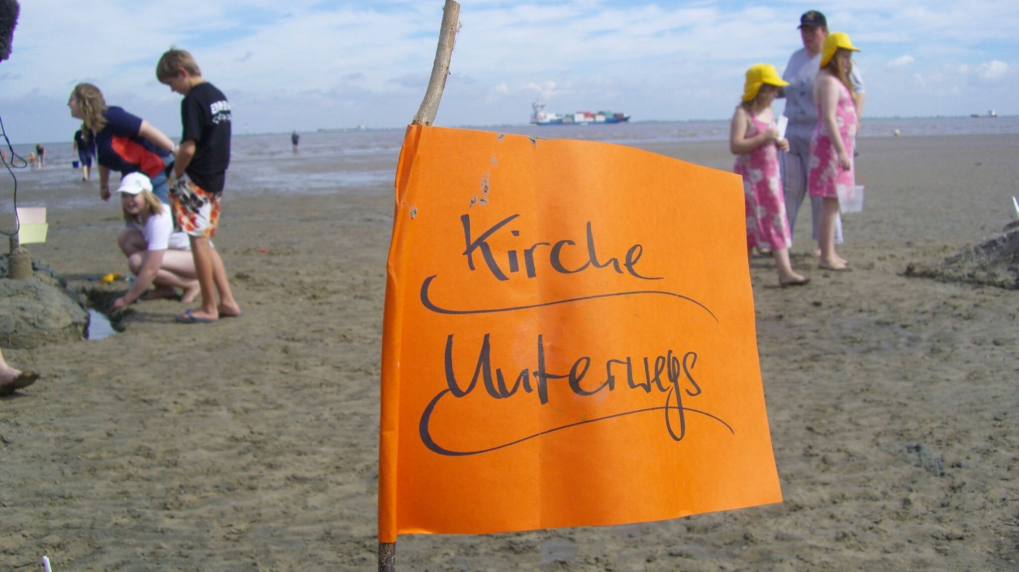 Eine orangefarbene Flagge steht auf einem Strand. Darauf steht: Kirche unterwegs. Im Hintergrund sieht man Menschen und das Meer.