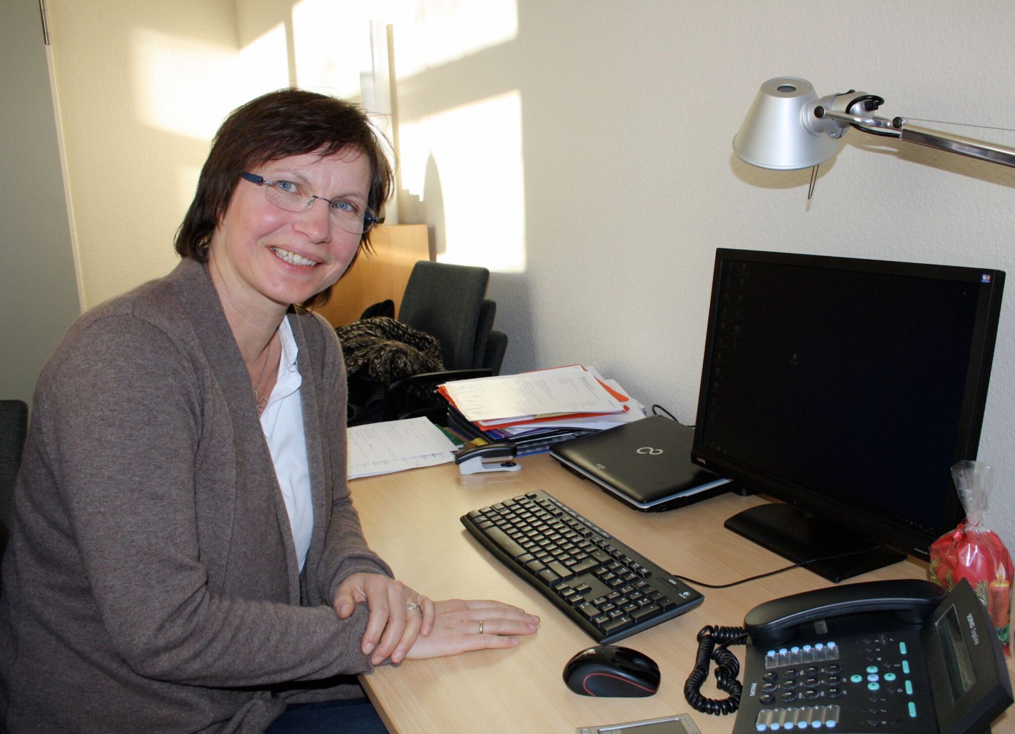 Eine Frau sitzt am Schreibtisch vor einem Computer. Sie trägt eine graue Strickjacke und eine Brille und hat braune, kurze Haare. Sie lächelt in die Kamera.