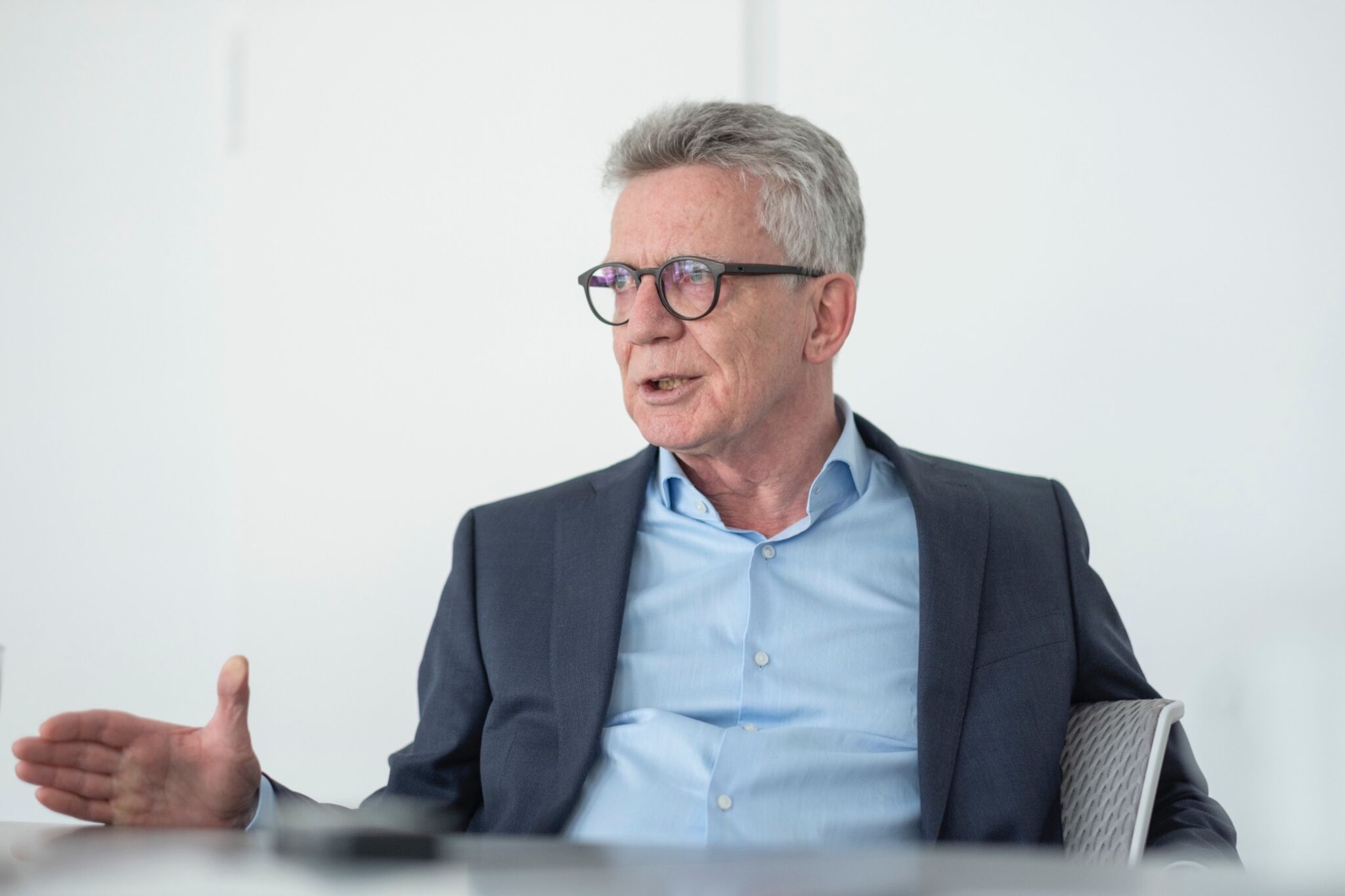 Das Bild zeigt Thomas de Maiziere. Er trägt einen grauen Anzug und ein hellblaues Hemd. Er hat graue Haare und trägt eine Brille. De Maiziere sitzt an einem Tisch und spricht.