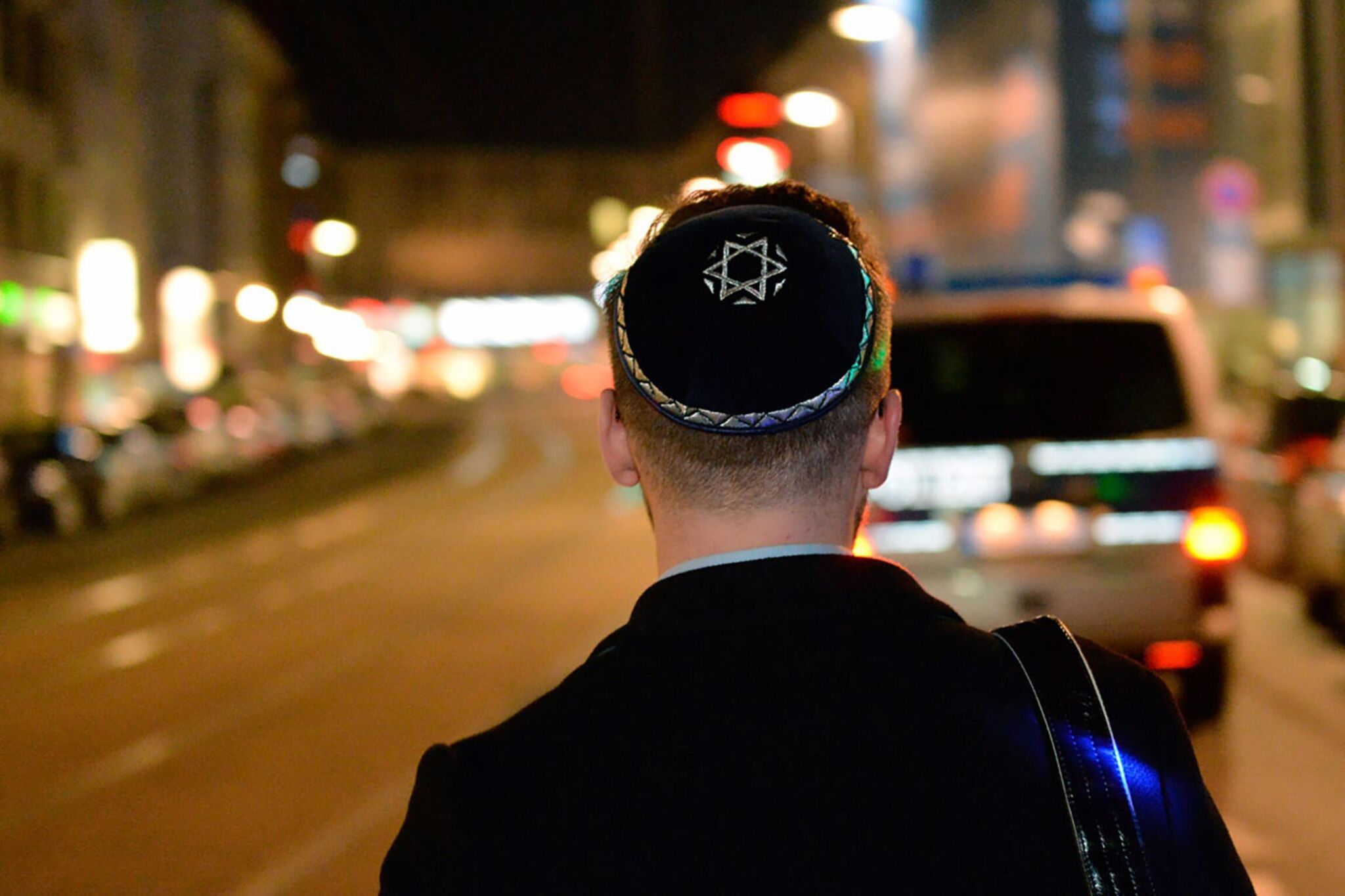 Ein Mann mit Kippa geht am Abend durch eine Stadt. Im Hintergrund sieht man ein Auto und Leuchtreklamen.