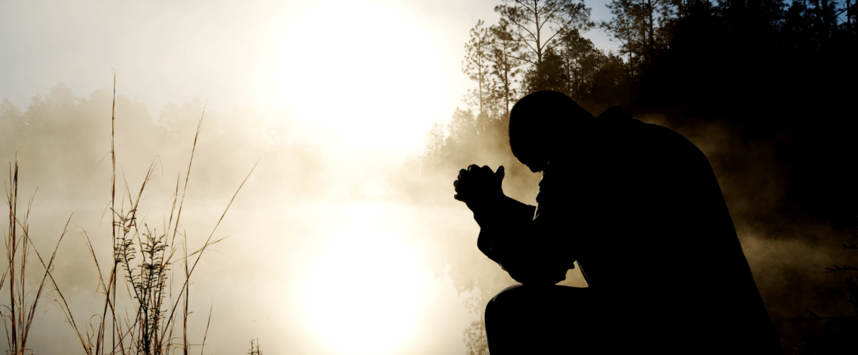 Eine nur im Schattenriss erkennbare Person sitzt an einem See, über dem Nebel liegt und faltet betend die Hände. Die Sonne durchscheint den Nebel mystisch.