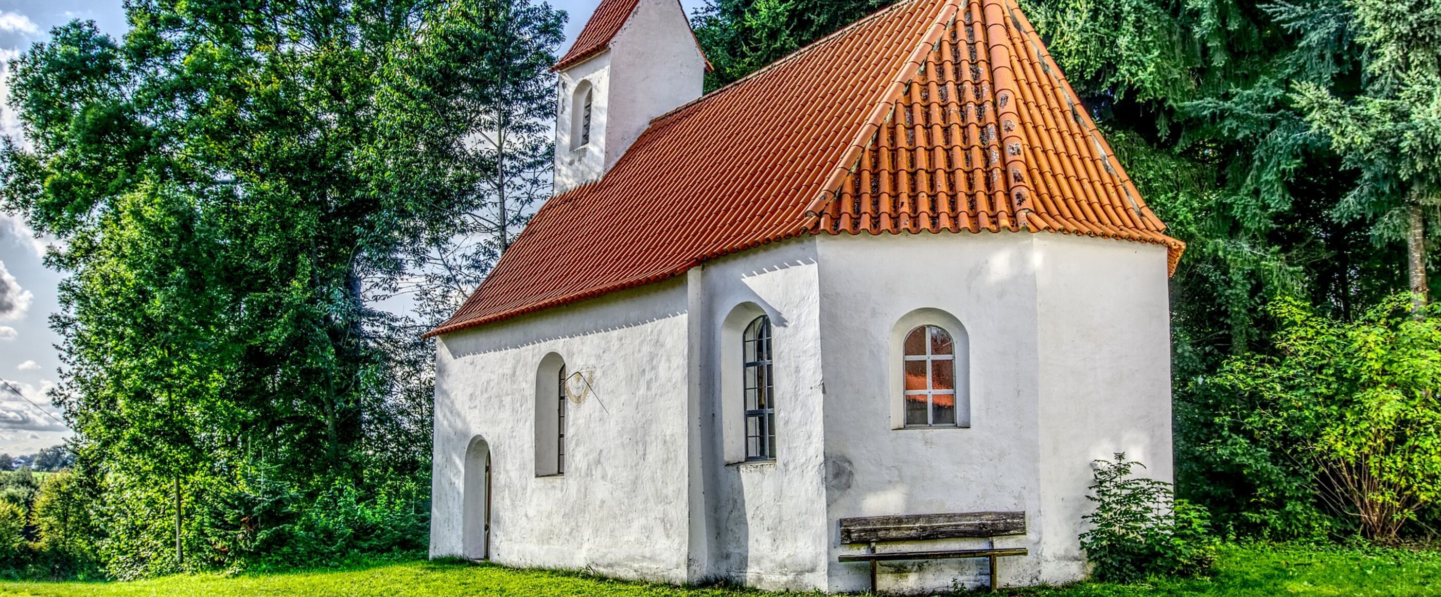 Eine Kapelle mit weißen Wänden und rotem Dach im Sonnenlicht; davor steht eine alte Holzbank.
