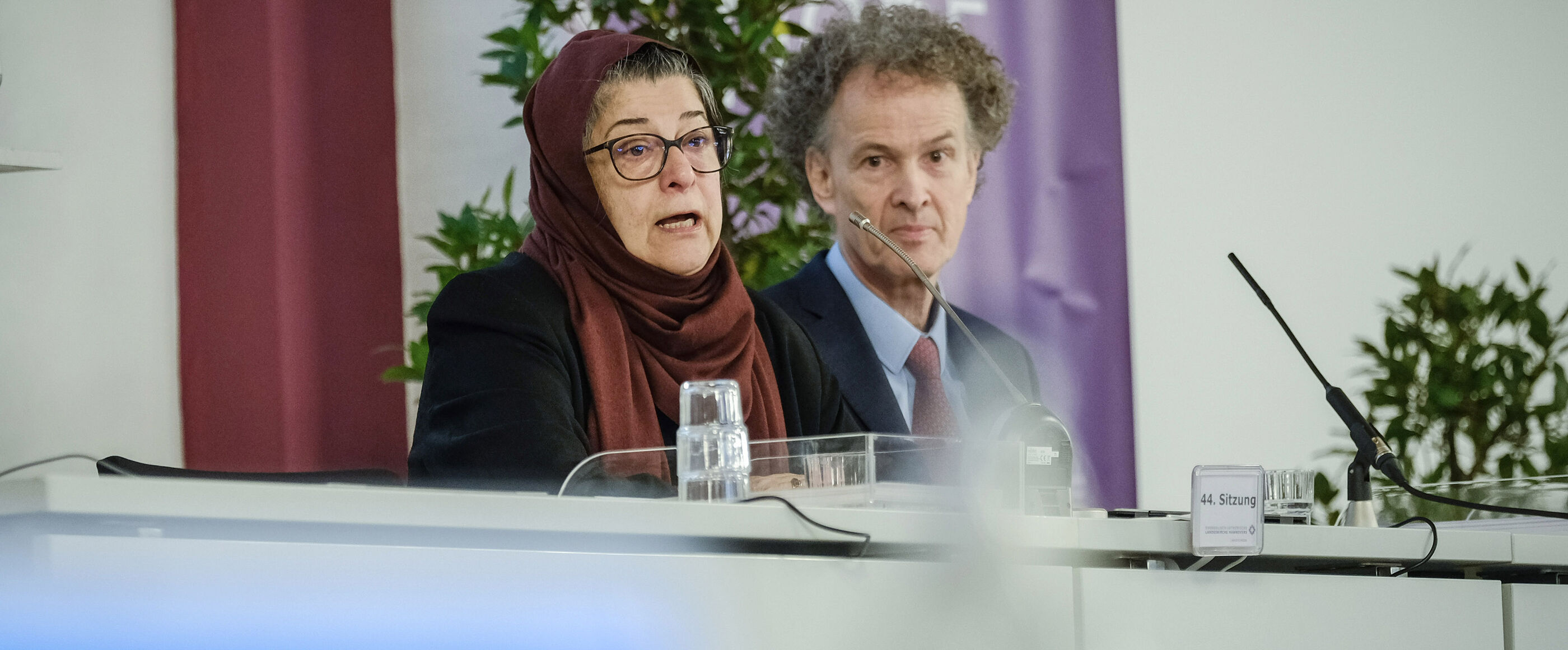 Eine weiblich lesbare Person mit islamischem Kopftuch, eine weiblich lesbare Person mit Schal und eine männlich lesbare Person im Anzug sitzen nebeneinander