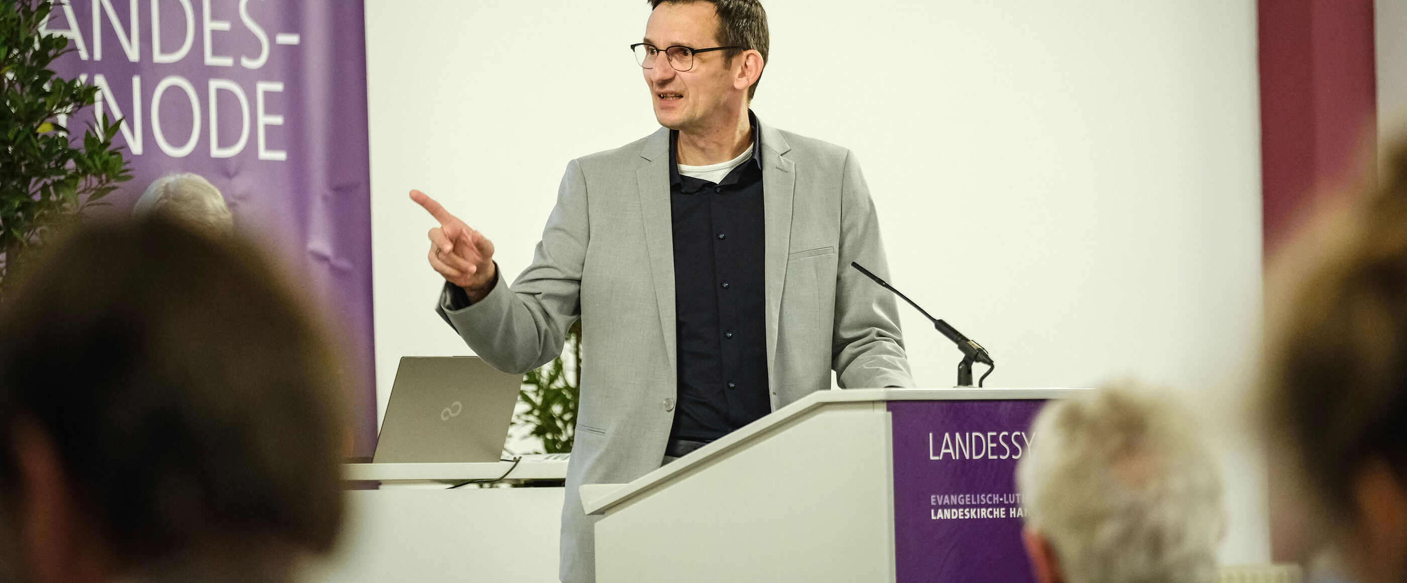 Eine männlich lesbare Person mit Brille und dunklen Haaren steht im Anzug hinter einem Rednerpult und spricht in ein Mikrofon.