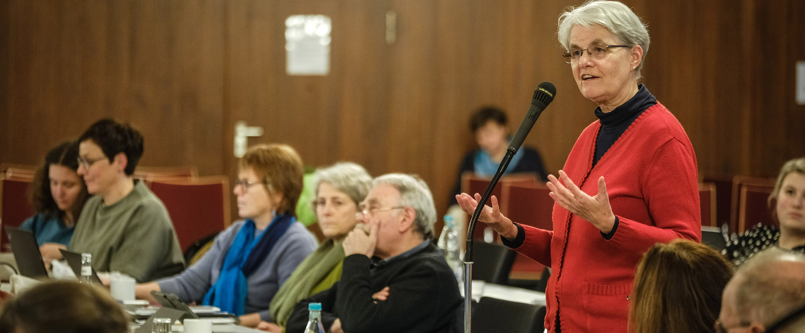 Eine weiblich lesbare Person mit Brille, kurzen blonden Haaren in einem roten Pullover steht im Plenum vor einem Mikrofon.