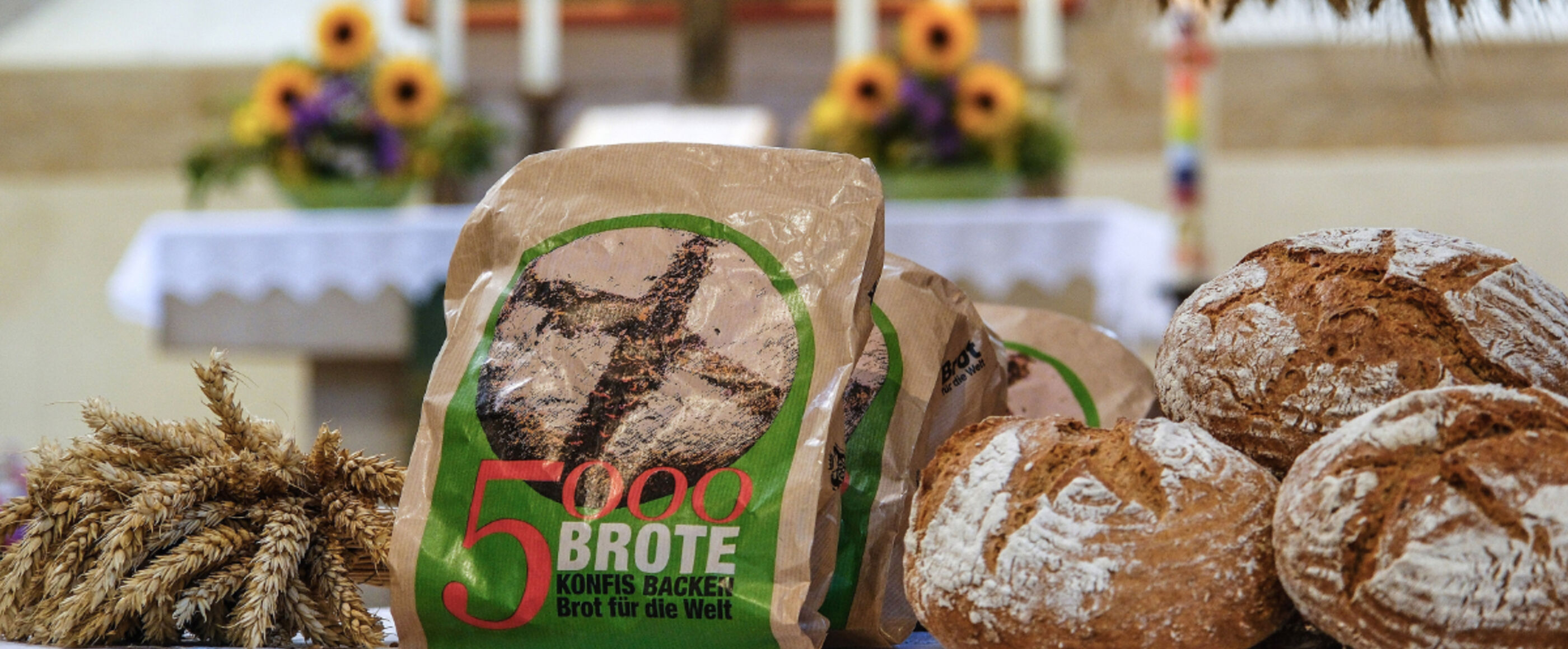 Man sieht frisch gebackene Brote. Sie liegen vor einem Altar in der Kirche. Die Kirche ist für das Erntedankfest geschmücke. In der Mitte der Brote stehen Brottüten mit dem Aufdruck: 5000 Brote - Konfis backen für Brot für die Welt.
