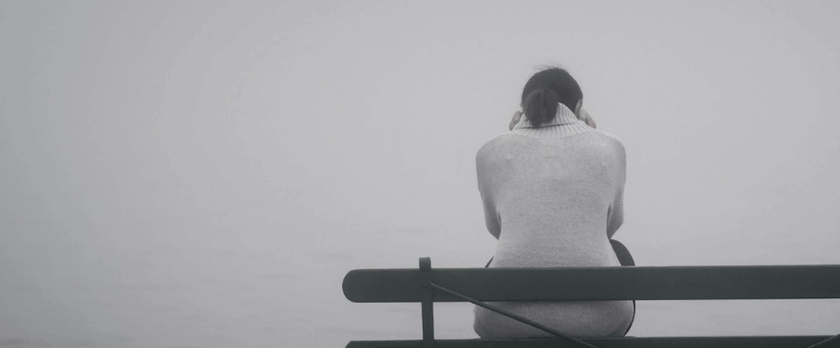 Eine junge Person hockt im Nebel auf einer Bank, alles ist grau.
