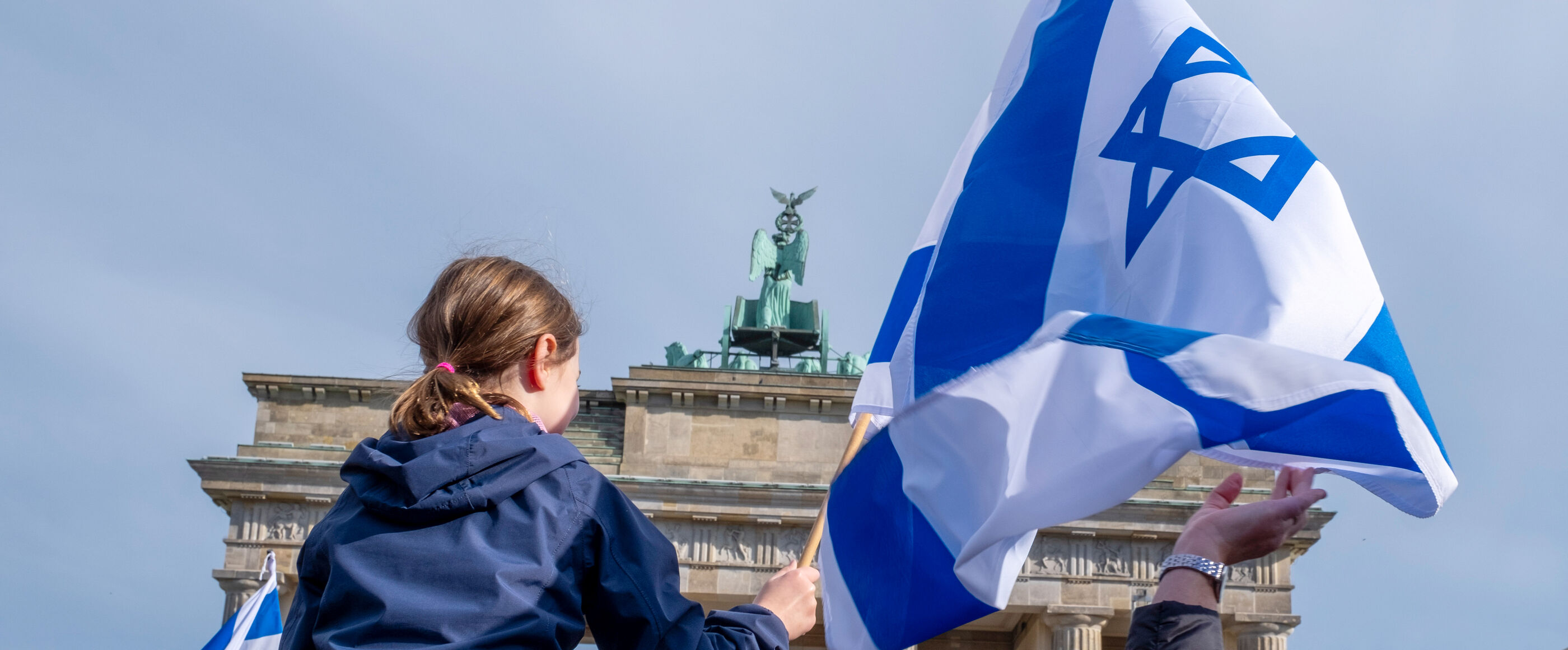 Im Hintergrund das Brandenburger Tor, im Vordergrund sitzt ein Kind auf den Schultern eines Menschen und hält eine Israel-Fahne.