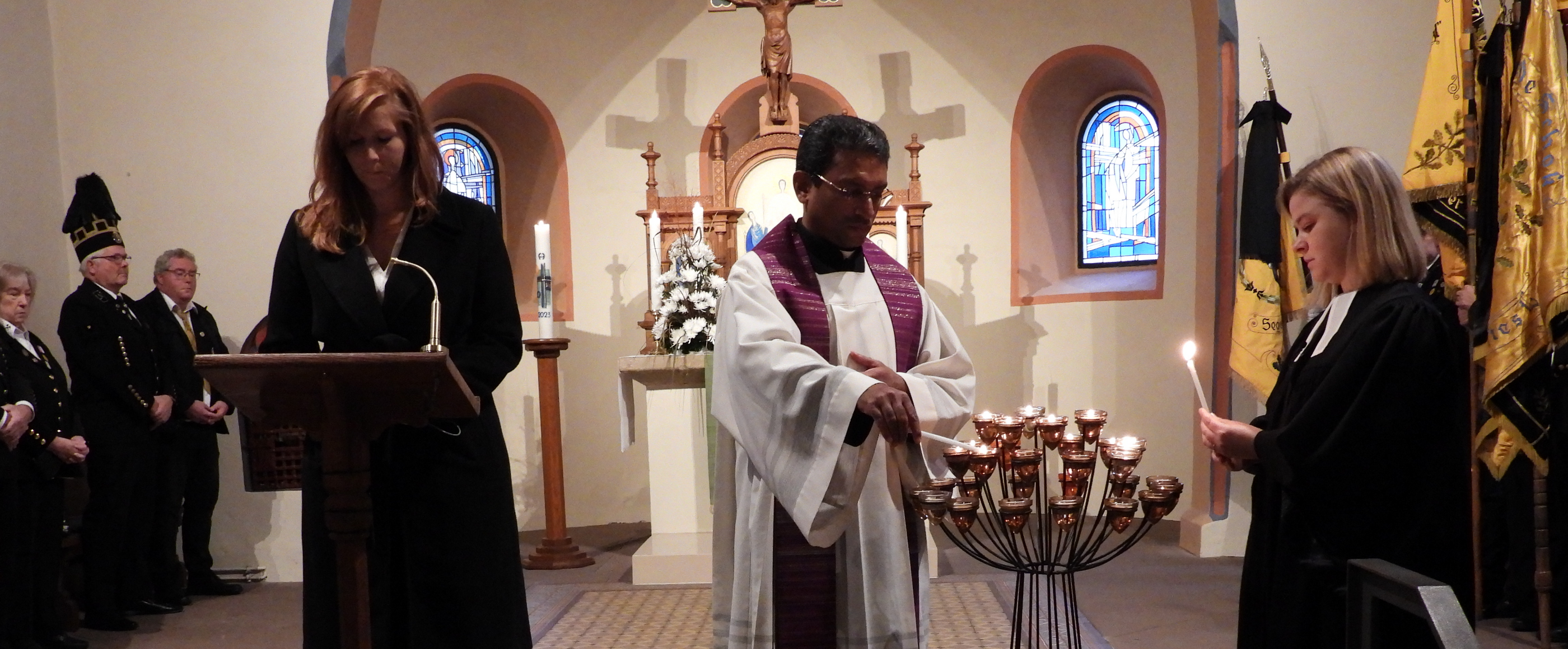 Eine Person steht an einem Pult in einer Kirche, daneben zündet ein Mann Kerzen an, daneben steht eine Frau und zündet ebenfalls Kerzen an.