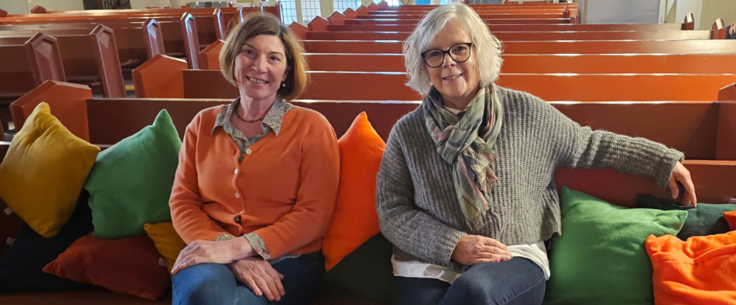 zwei Frauen sitzen auf Bänken mit bunten Sitzkissen in einer Kirche