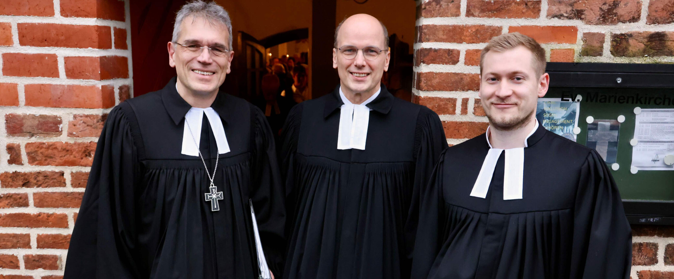 Drei Männer in Talar stehen vor einer Kirche und lächeln in die Kamera.