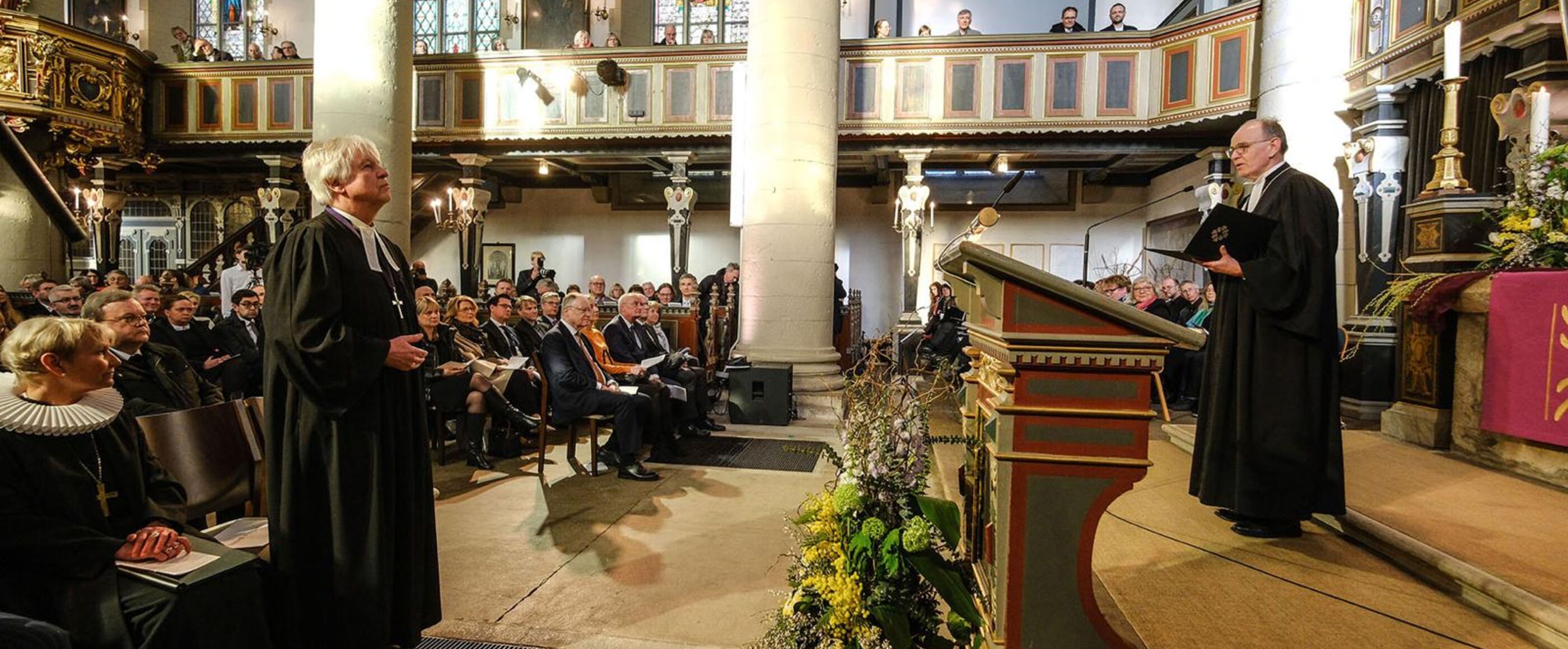 Zwei männlich zu lesende Personen im Taler stehen sich im Altarraum einer Kirche gegenüber.