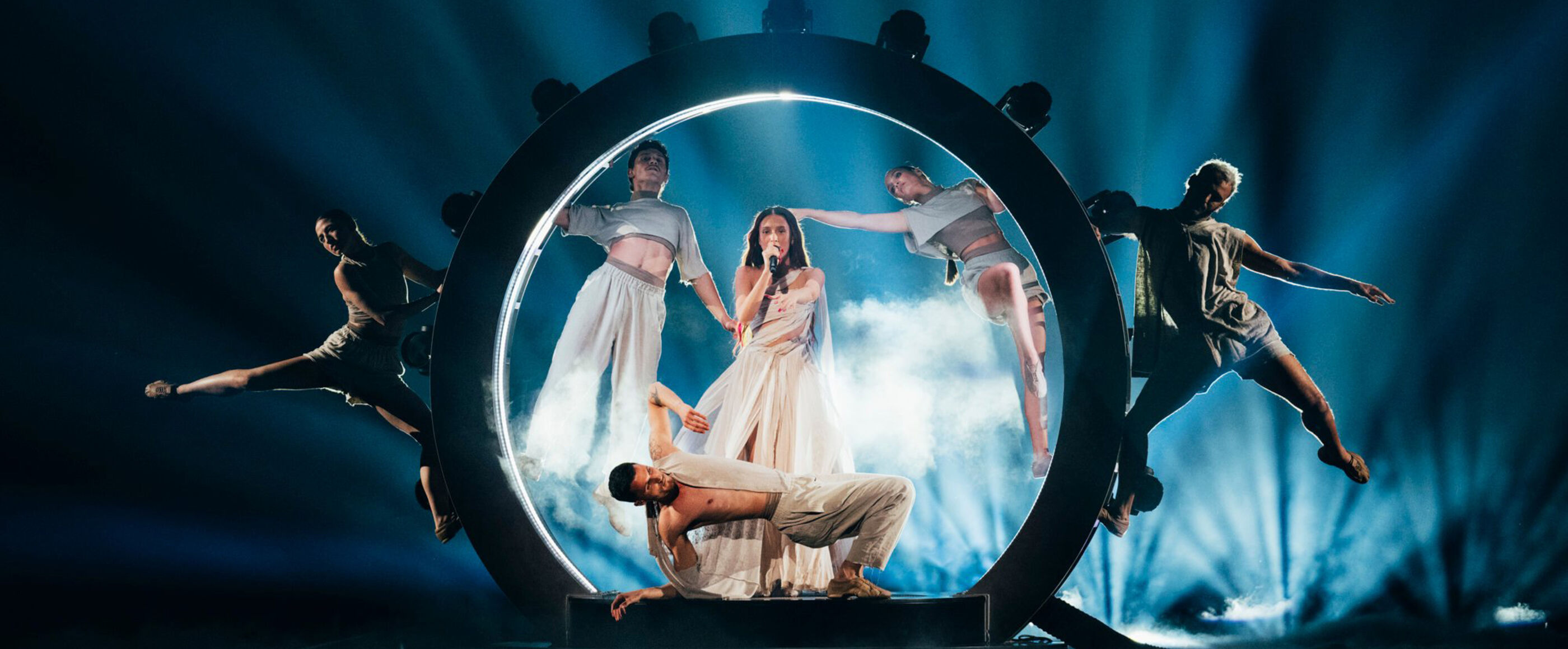 Eine als Frau lesbare Person singt in einem beleuchteten Kreis, umgeben von Tänzern.