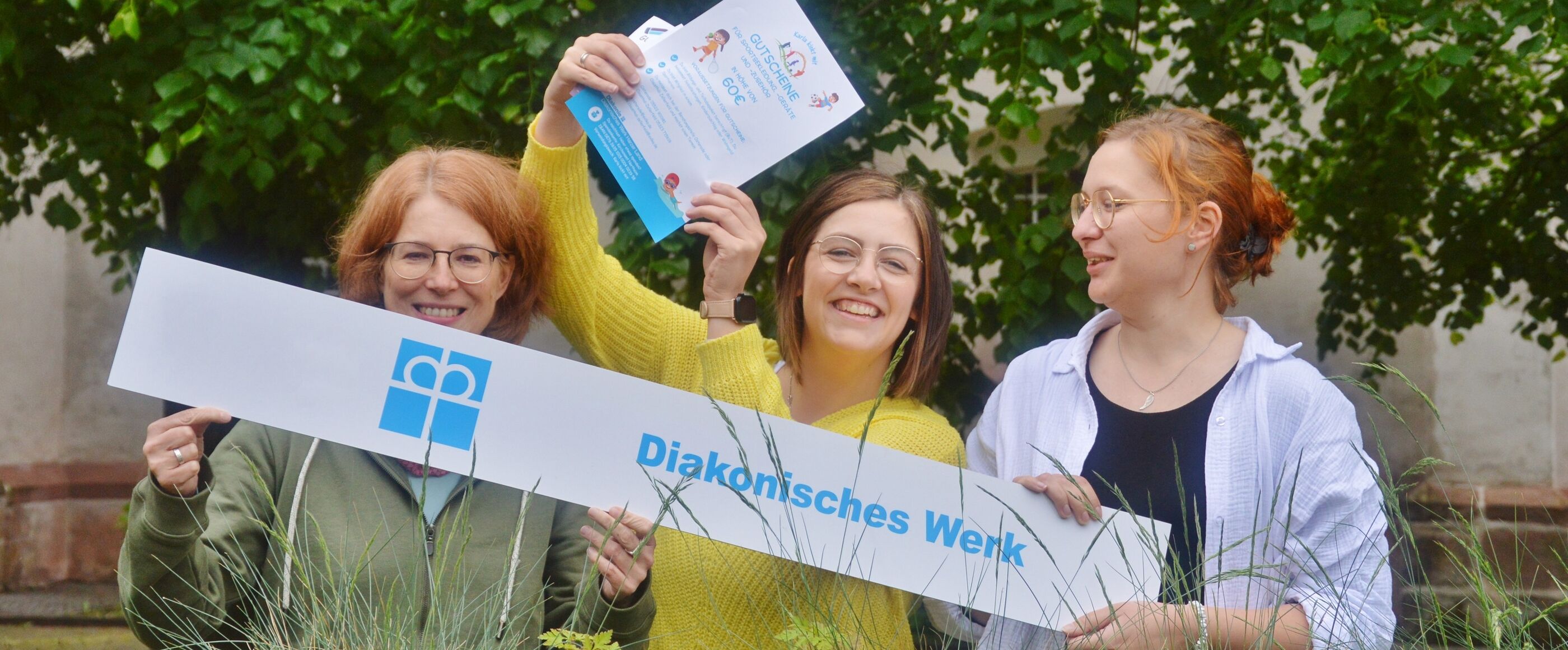 Ein Foto von drei weiblich gelesenen Personen, die in die Kamera lächeln. Sie halten ein weißes Banner mit blauer Schrift "Diakonisches Werk". Die mittlere Person hält eine Urkunde. Sie stehen im Grünen. 