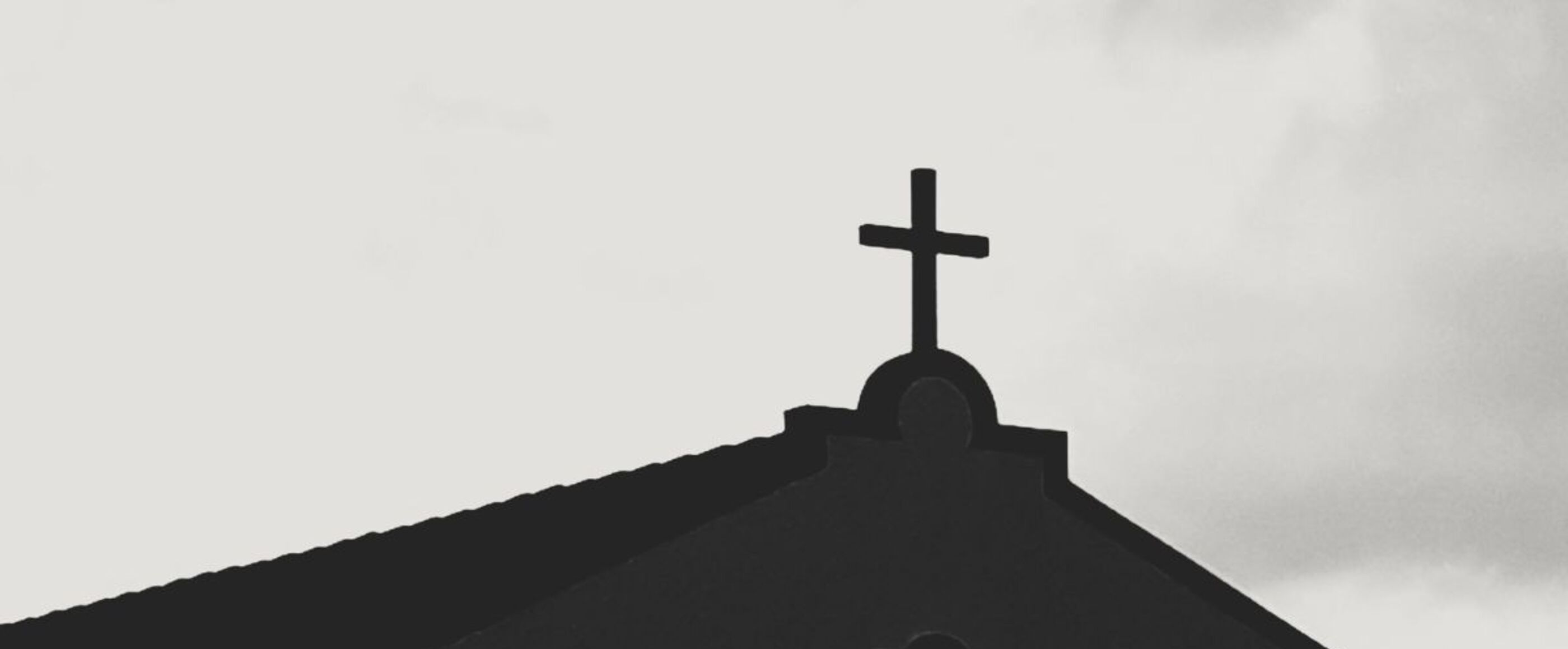 Gegen einen grauen Himmel zeichnet sich auf einem Spitzdach ein Kreuz ab.
