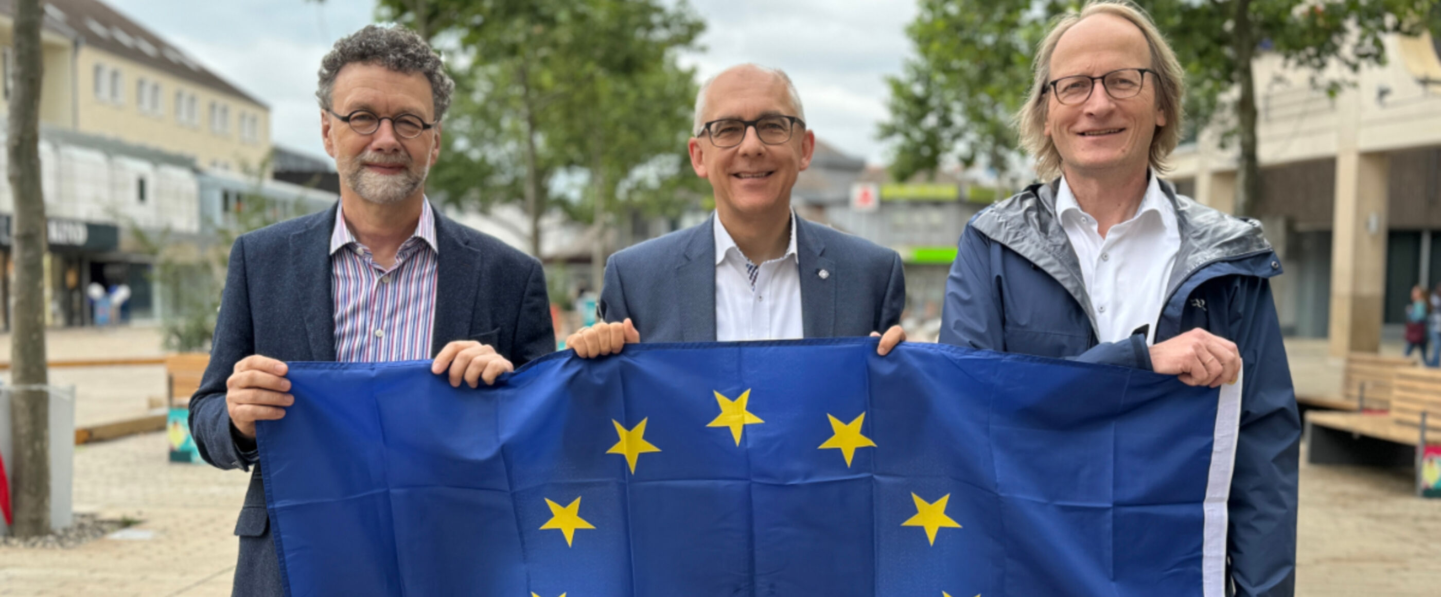 drei männlich gelesene Personen halten eine Europaflagge in der Hand.