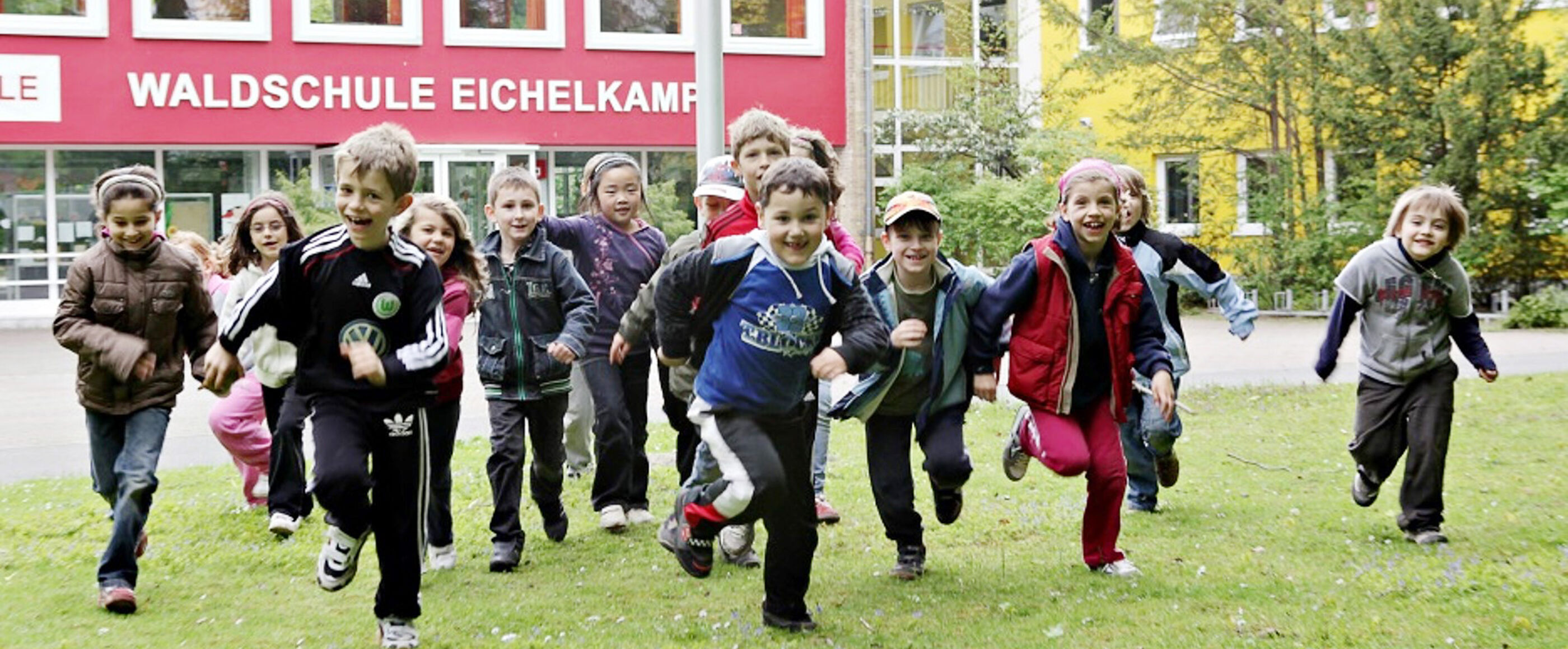 Kinder laufen vor einem roten Schulgebäude auf die Kamera zu