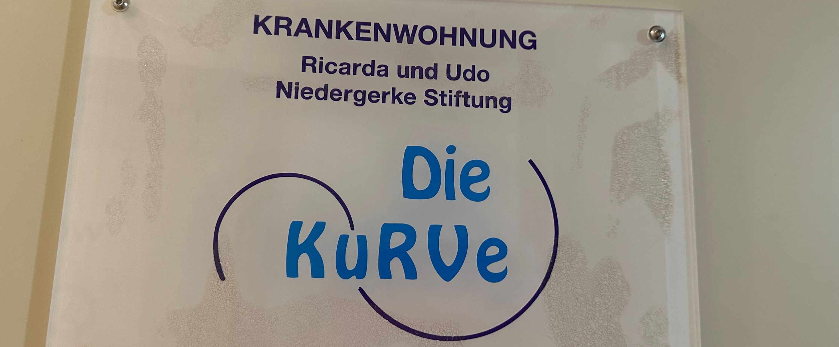 Ein Schild an der Wand, in blauer Schrift steht die Kurve drauf. Darüber in schwarzer Schrift: Krankenwohnung, Ricard und Udo Niedergerke Stiftung. 