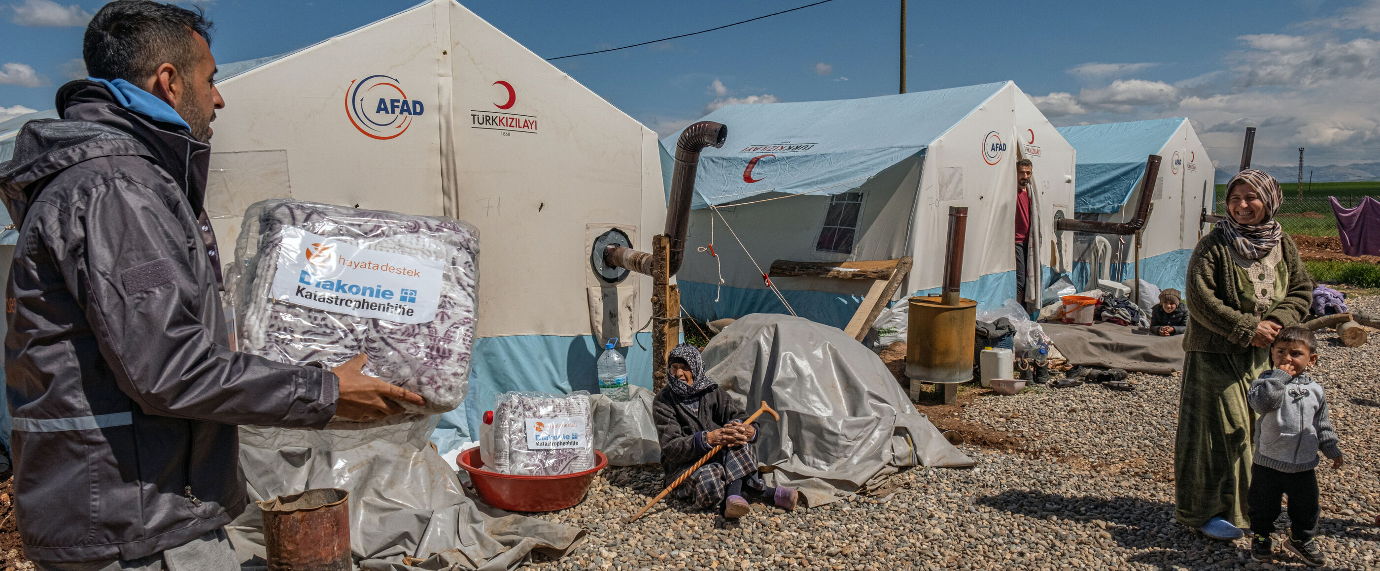 Eine männlich gelesene Person trägt ein Paket, auf dem "Diakonie Katastrophenhilfe" steht, zu einer weiblich gelesenen Person mit Kind. Das Umfeld wirkt wie in einem Flüchtlingslager: Zelte, Kiesboden.