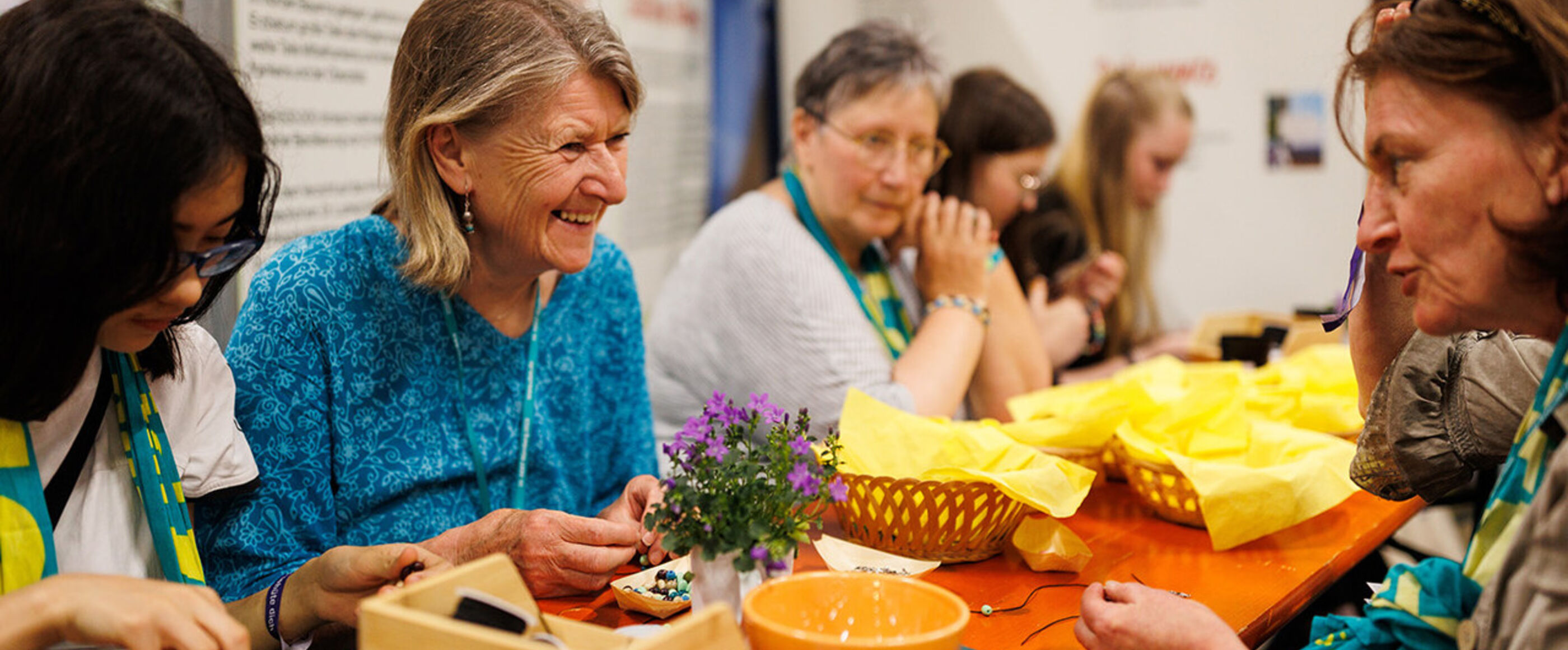 Weiblich gelesene Personen unterschiedlichen Alters sitzen an einem langen Tisch und knüpft Perlenarmbänder, sie tragen den Kirchentagsschal aus Nürnberg, manche unterhalten sich und lachen