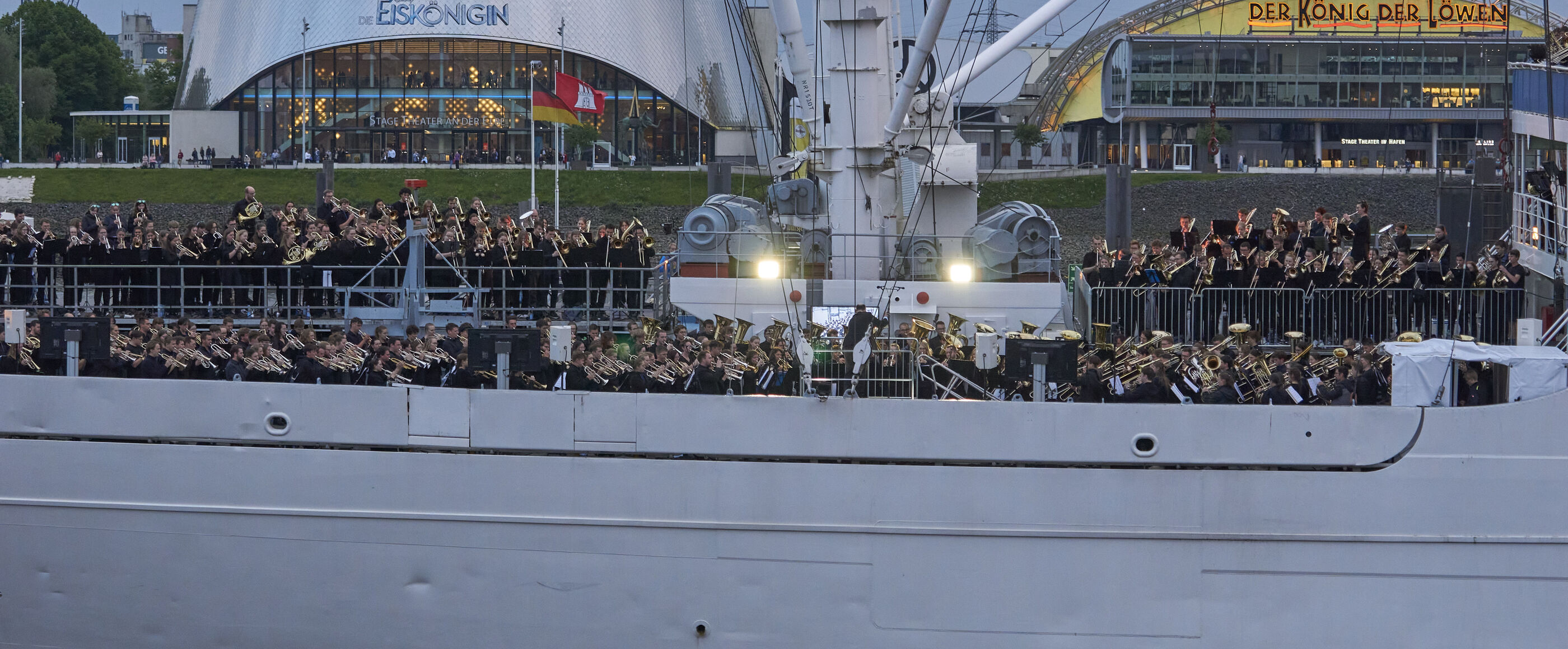 Blick auf die Seite eines Schiffs im Wasser. Das Deck ist sehr gut gefüllt mit Menschen, die teils Instrumente in Händen halten. 