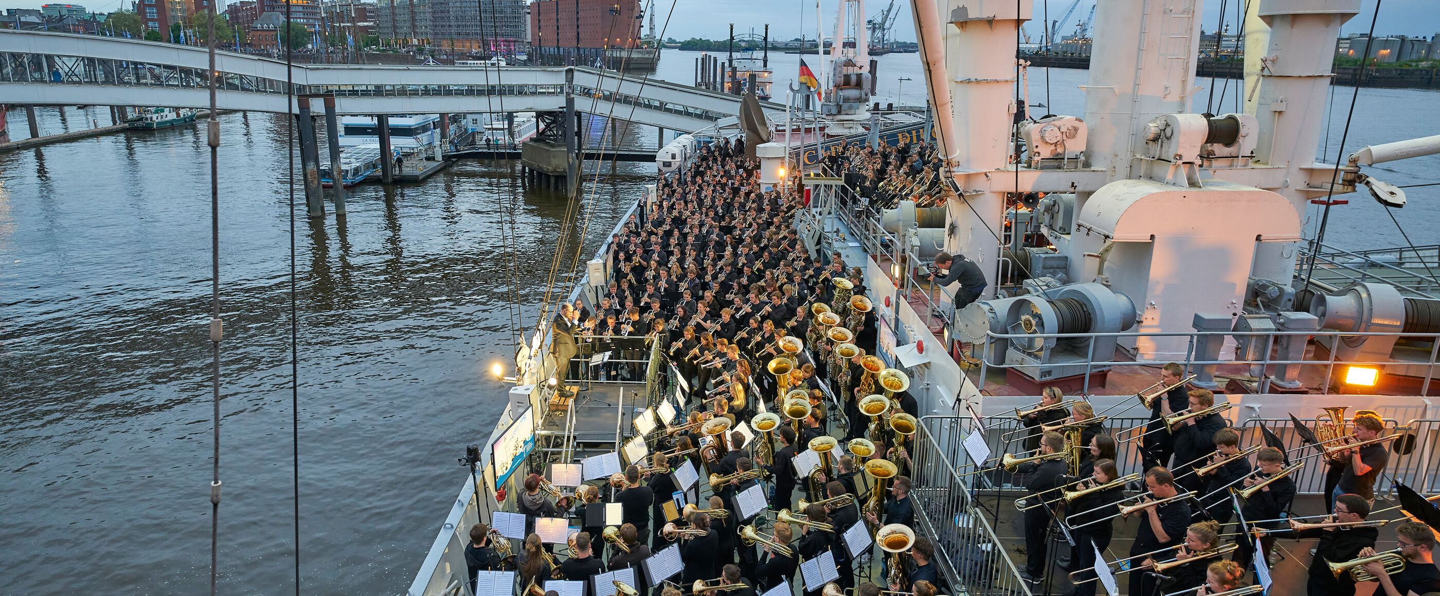 Ein riesige Blasensemble spielt auf einem Schiff, im Hintergrund ist die Hamburger Elbphilharmonie zu sehen.