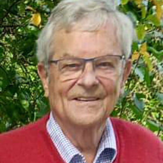 Ein älterer Mann mit grauen Haaren und Brille. Er trägt einen roten Pullover über dem Hemd.
