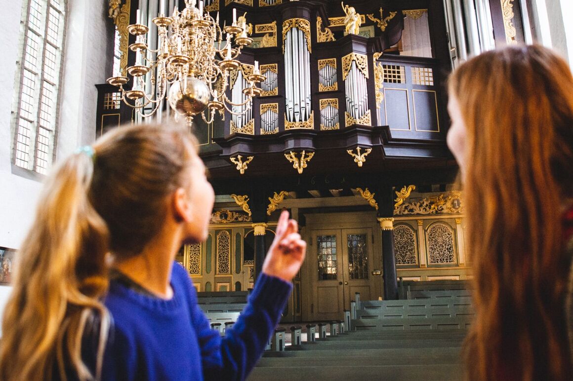 Zwei weiblich gelesene Personen stehen in einer Kirche und deuten auf eine Orgel